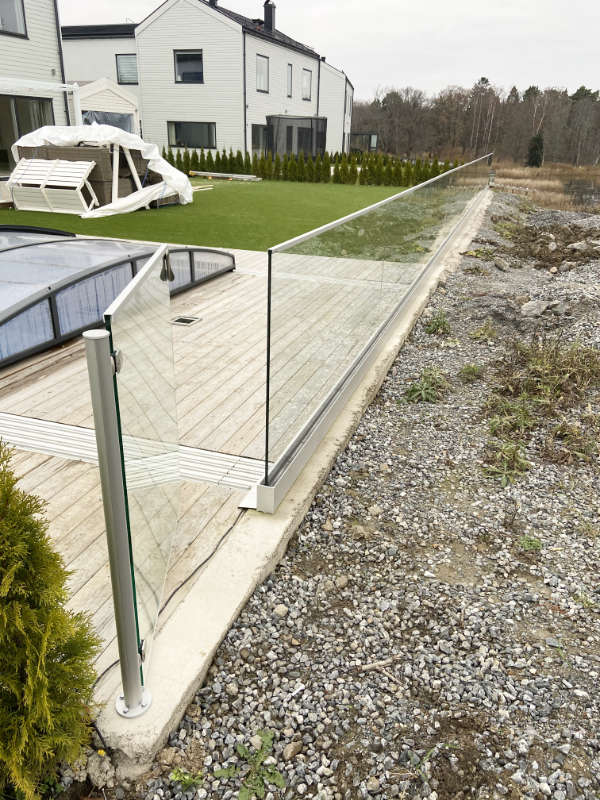 Glasräcke utomhus på trädäck, fäst med en silverfärgad basprofil och en stolpe.