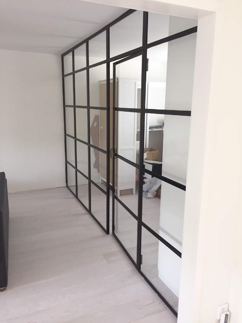 Modern rumsavdelare med svart stålram och glasdörr i en ljus hemmiljö.