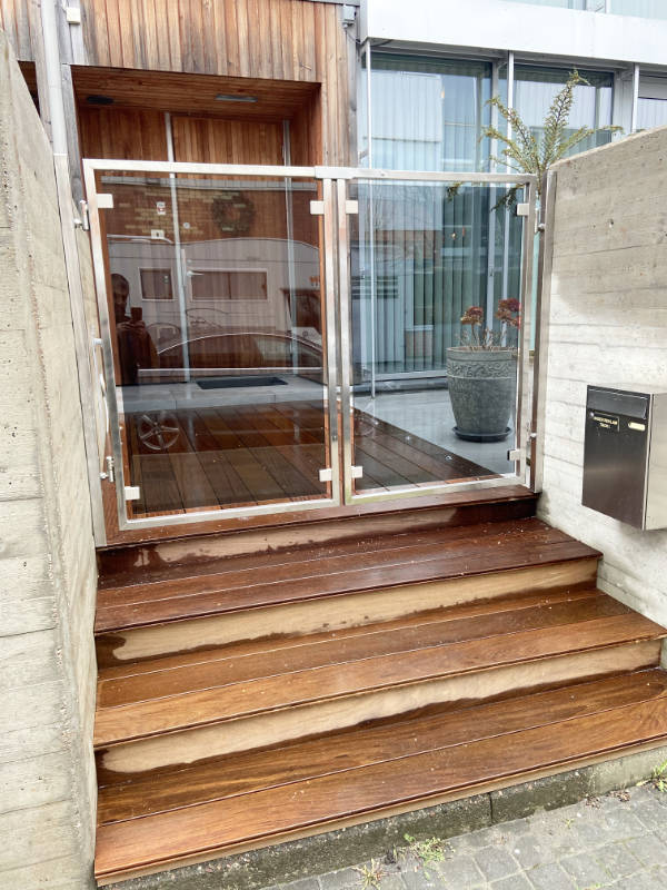 Trätrappa upp till en entré med en dubbel grind i glas och rostfritt stål mellan betongväggar.