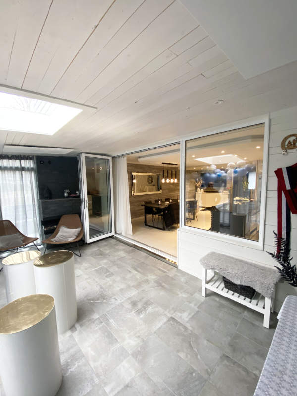 Ljust uterum med grått klinkergolv, vikbara glasdörrar och vita sidobord med guldfärgade toppar.