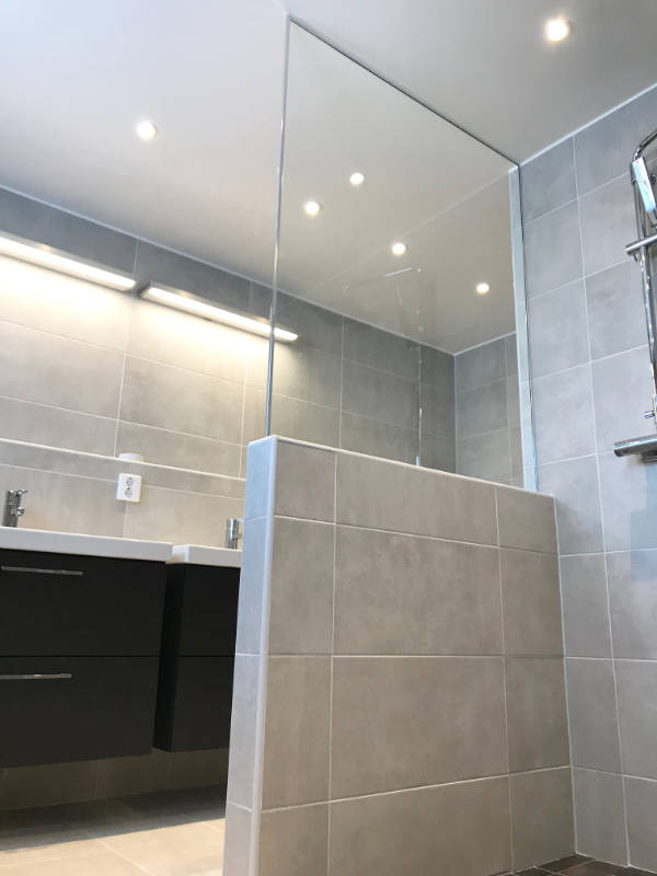 Duschvägg i klarglas ovanpå en låg kaklad vägg i ett grått, modernt badrum.